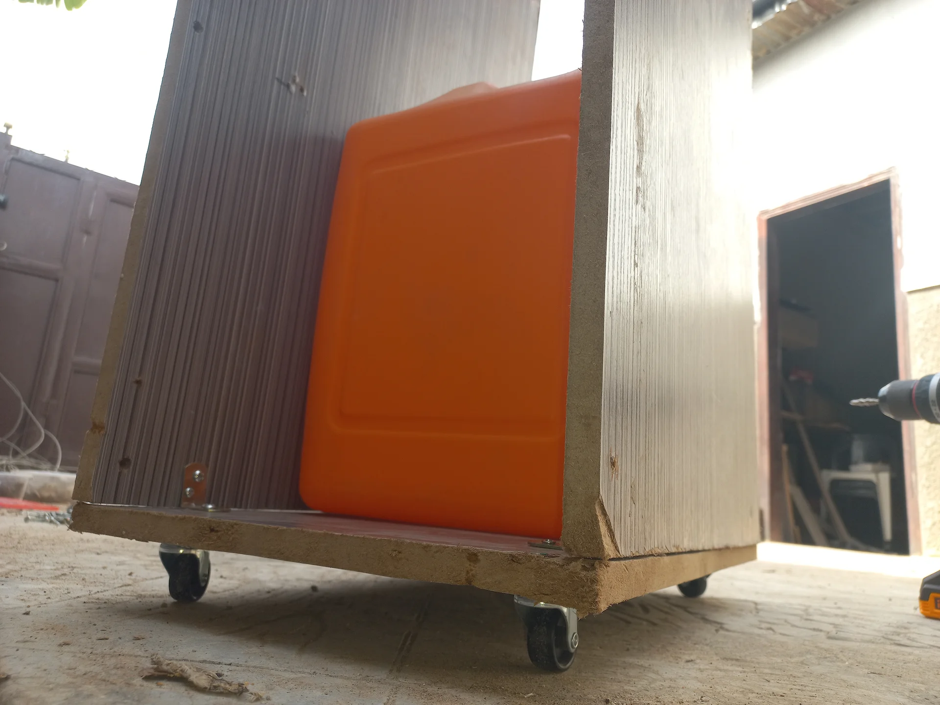 The bin frame on four castor wheels with corrugated side panels and an orange front panel already fitted, photographed from a low angle outdoors
