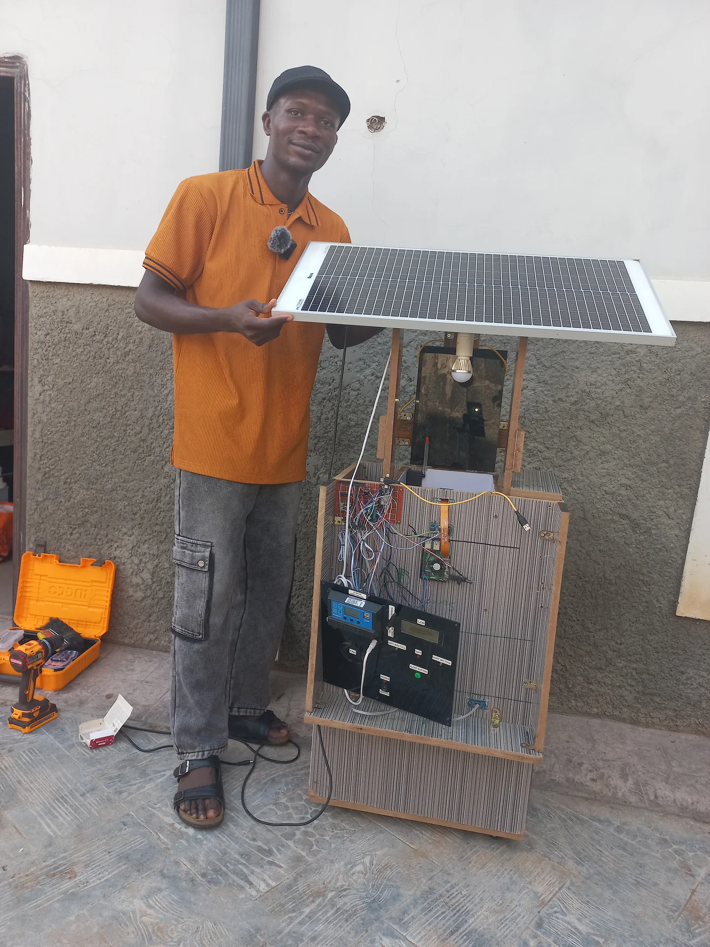 Builder standing next to the assembled system, steadying the solar panel already mounted on the overhead frame, with the electronics panel face visible at front