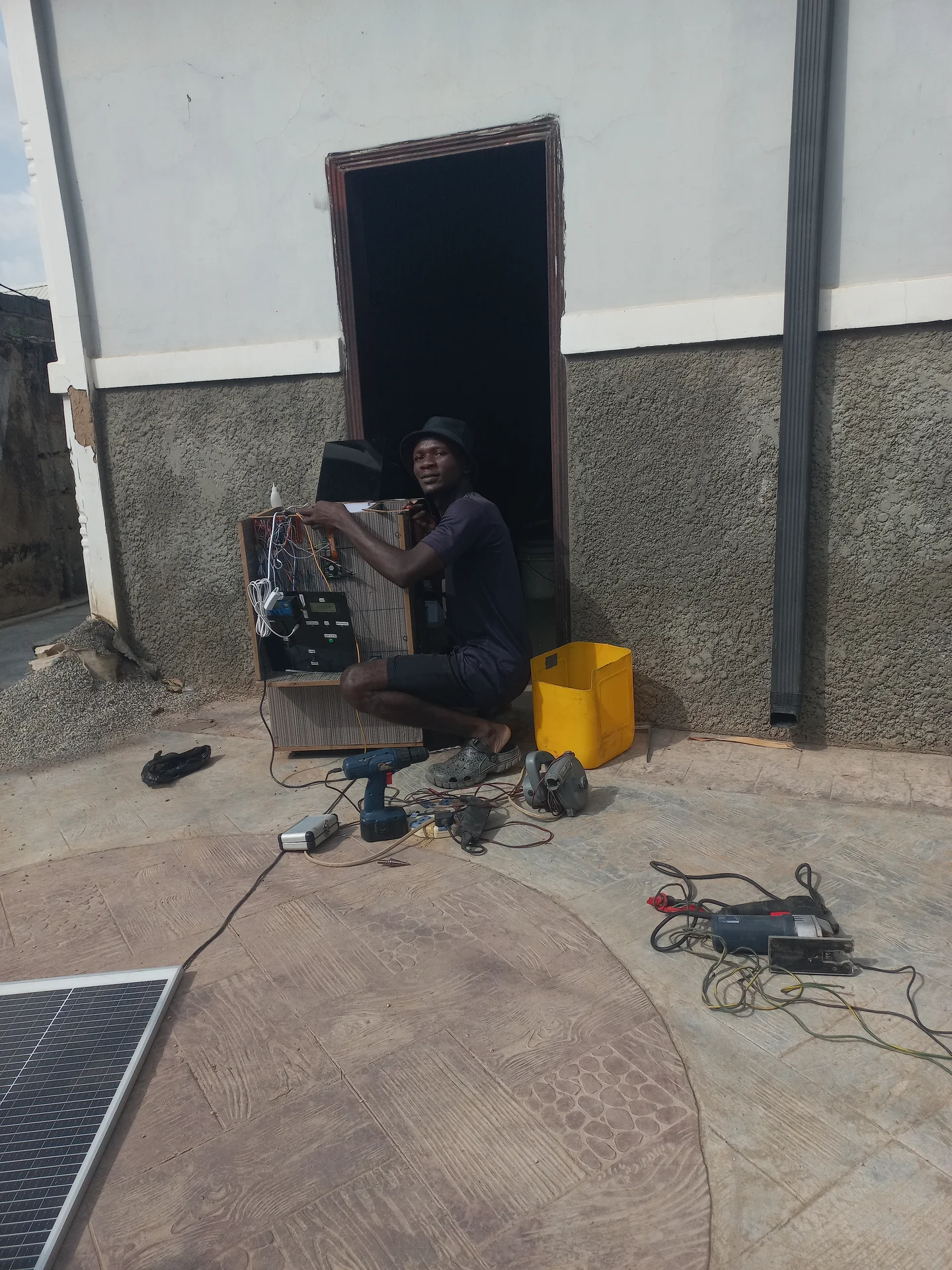 Builder crouching beside the bin outdoors, working on the open electronics bay, with the solar panel on the ground in the foreground