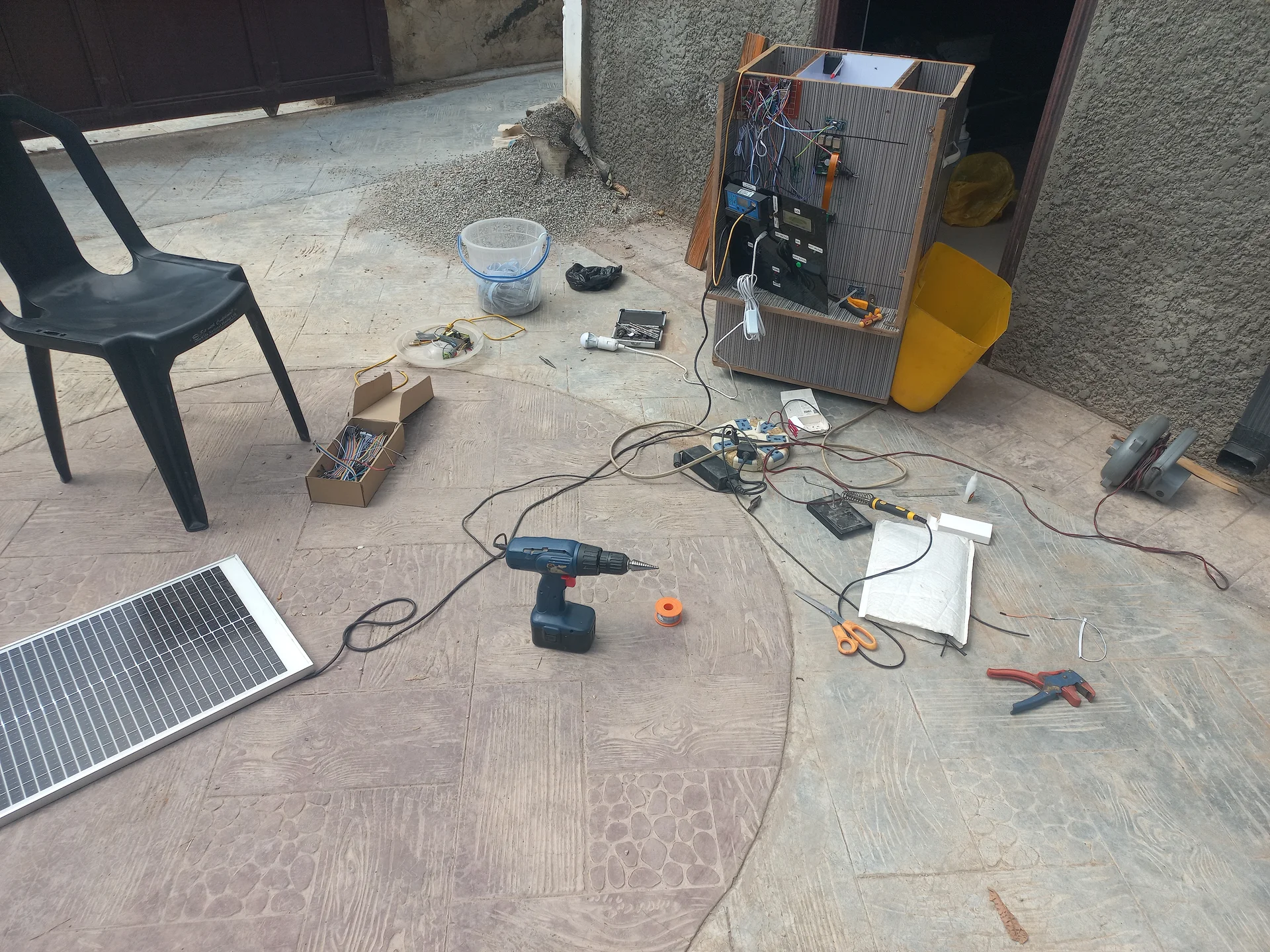 Wide outdoor shot showing the full workshop scene with the bin sitting on the ground, the electronics bay exposed, the solar panel and tools scattered around