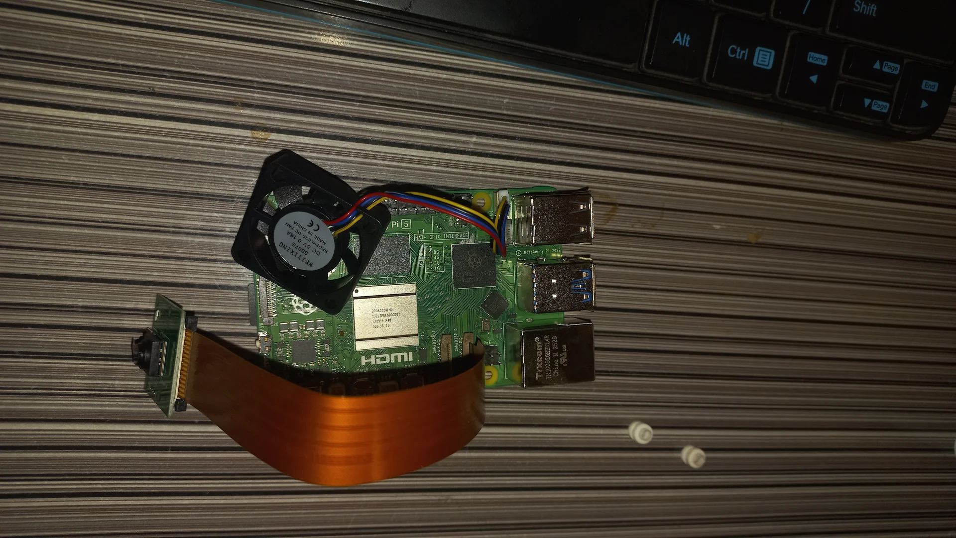 Raspberry Pi 5 with a small blower fan placed on the processor and the Pi Camera Module with ribbon cable laid alongside, before final assembly