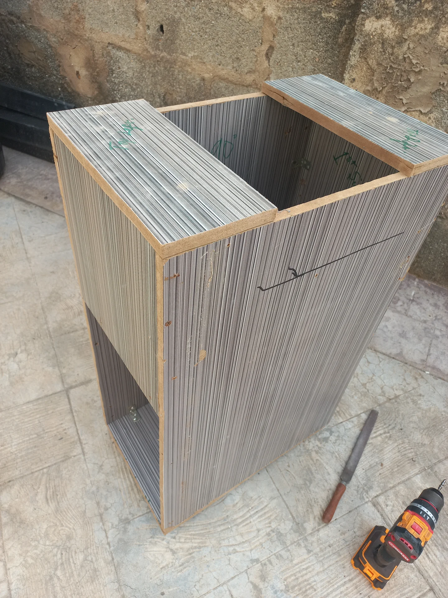 Three-quarter angled view of the bin with the front panel not yet fitted, revealing the interior compartment divider and a lower opening, with tools on the ground