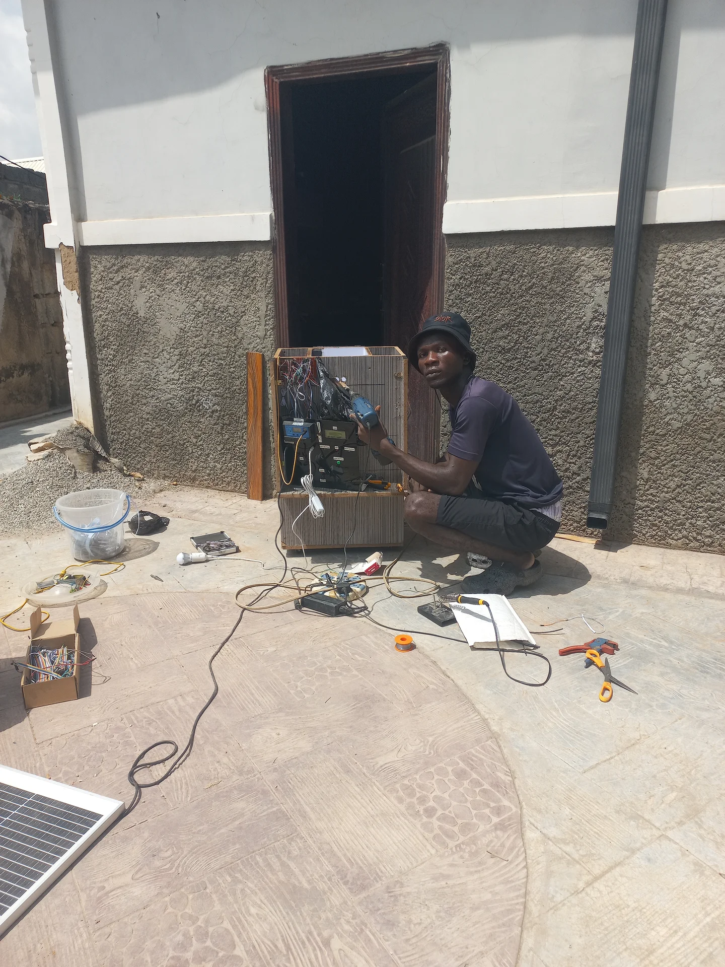 Builder crouching outdoors while working on the open electronics bay of the bin, with tools and the solar panel laid flat on the ground around him