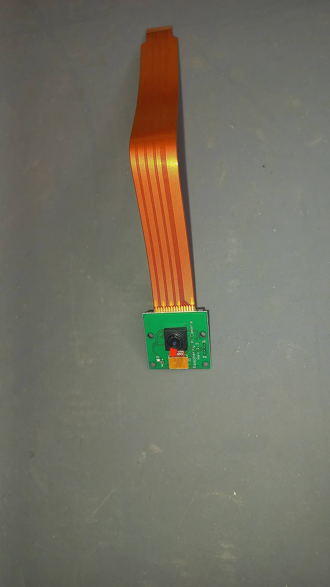 Pi Camera Module v2 lying flat with its orange CSI ribbon cable attached, showing the lens, red LED and PCB labeling